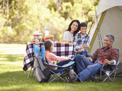 A family of four enjoys a picnic by their tent in a sunny, green park. They are smiling and relaxed, with food and drinks on a plaid tablecloth.